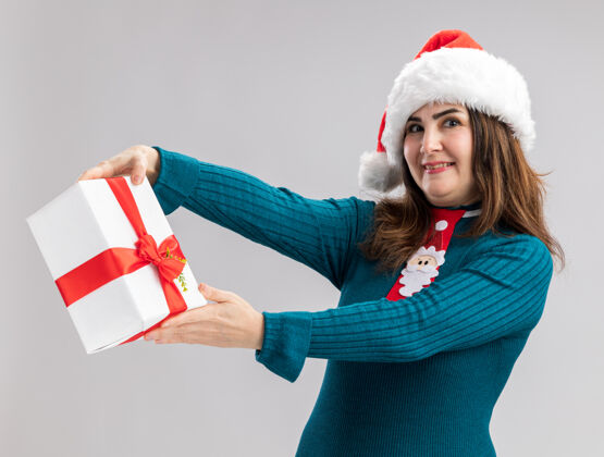 高兴戴着圣诞帽 系着圣诞领带的成年白种女人拿着圣诞礼品盒 看着相机 在白色背景上 有着独立的复制空间成人圣诞老人举行