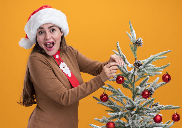 就在附近兴奋的年轻漂亮女孩戴着圣诞帽 打着领带 站在橙色墙上的圣诞树旁圣诞节领带帽子