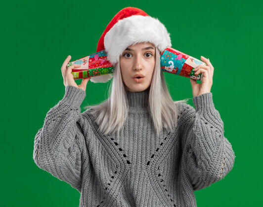 困惑年轻的金发女郎 穿着冬衣 戴着圣诞帽 耳朵上戴着五颜六色的纸杯 站在绿色的背景下 看起来很困惑毛衣站着年轻