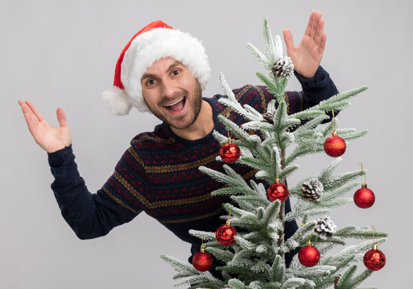 年轻令人印象深刻的年轻白人男子戴着圣诞帽站在圣诞树后 保持双手在空气中隔离在白墙上圣诞节印象深刻男人