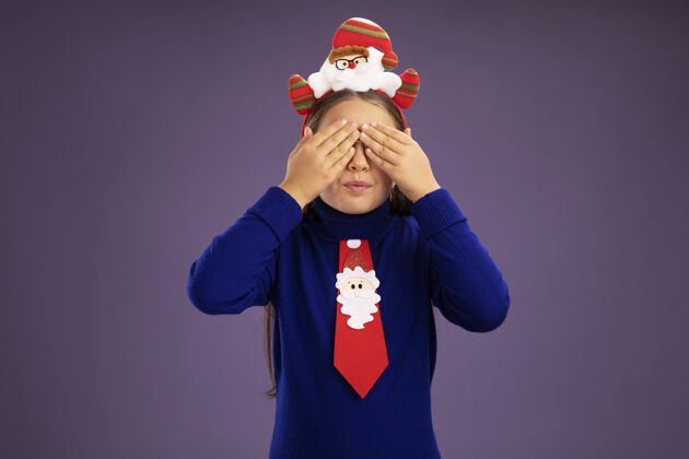 边缘小女孩穿着蓝色高领毛衣 系着红色领带 头上戴着有趣的圣诞戒指 手放在紫色背景上遮住眼睛圣诞节头封面