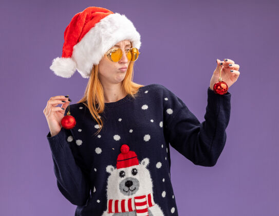 举行年轻漂亮的女孩穿着圣诞毛衣 戴着圣诞帽 戴着眼镜 拿着圣诞球看着紫色背景上的孤立球看眼镜