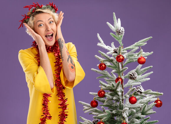 头兴奋的金发女郎头戴圣诞花环 脖子上戴着金箔花环 站在装饰好的圣诞树旁 手放在脸上 在紫色的墙上显得与世隔绝周围保持女人