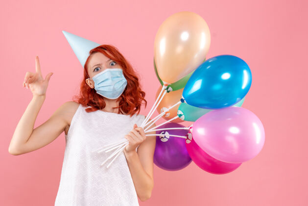 庆典前视图中的年轻女子拿着五颜六色的气球在无菌面具上粉红色的墙壁女性气球病毒