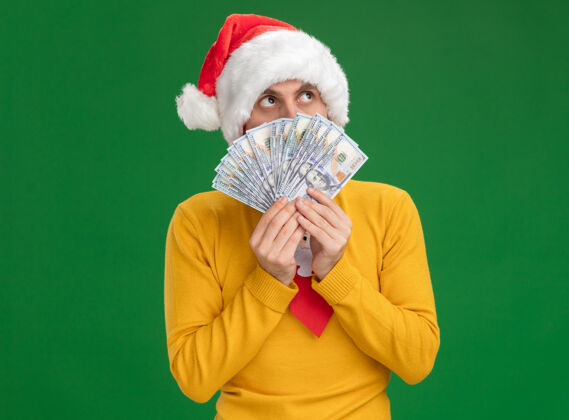 领带印象深刻的年轻白种人戴着圣诞帽 打着领带 手里拿着钱 从后面抬头望去 隔离在绿色的墙上 留着复印空间男人圣诞节帽子