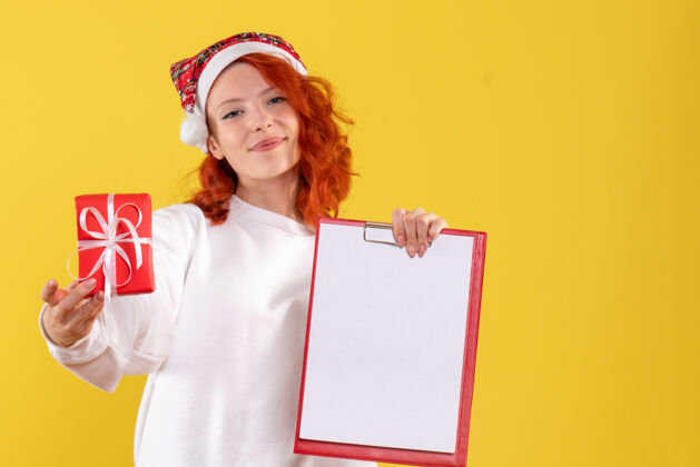 节日手持小圣诞礼物和黄色墙上的文件笔记的年轻女子的正面视图便条礼物成人