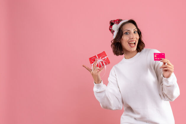 年轻年轻女子手持小圣诞礼物和粉红色墙上的银行卡的正面视图年轻的女性女圣诞节