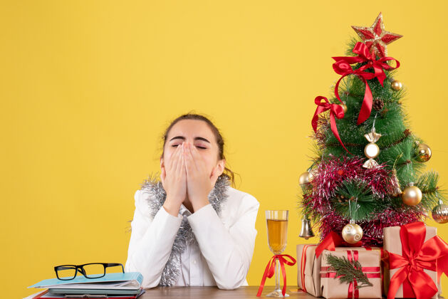 生意前视图：女医生坐在桌子后面 拿着圣诞礼物 黄色背景上的树打着哈欠打哈欠新的情感