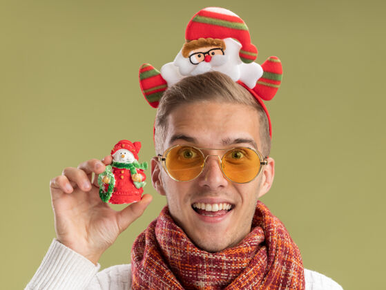 快乐特写镜头给人留下深刻印象的年轻帅哥戴着圣诞老人的头带和围巾看着相机拿着雪人圣诞饰品孤立在橄榄绿的背景雪人圣诞快乐圣诞老人