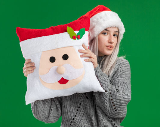 脸快乐的金发女郎 穿着冬衣 戴着圣诞帽 抱着圣诞枕头 面带微笑地看着相机 站在绿色的背景下圣诞老人毛衣圣诞节