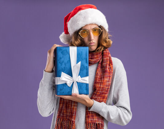 礼物戴着圣诞老人帽 戴着黄色眼镜 脖子上围着暖和的围巾的年轻人拿着圣诞礼物 严肃的脸站在紫色的背景上看着摄像机严肃站着年轻人