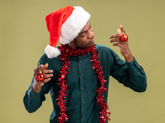 困惑一个戴着圣诞帽 戴着花环 拿着圣诞球的非裔美国人 站在绿色的背景下 满脸疑惑非洲怀疑圣诞节