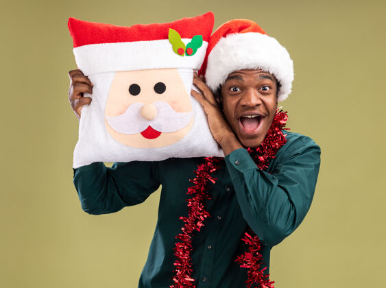 圣诞老人快乐和兴奋的非洲裔美国人戴着圣诞帽 戴着花环 抱着圣诞枕头 看着站在绿色背景上的相机抱着兴奋美国人
