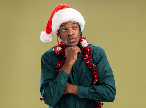 沉思快乐的非洲裔美国人戴着圣诞帽 戴着花环 耳朵上戴着圣诞球 站在绿色的背景上 带着沉思的表情望向一边花环舞会美国人