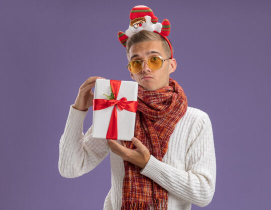 围巾困惑的年轻帅哥戴着圣诞老人的头带和围巾拿着礼包孤立在紫色的墙上圣诞老人年轻包