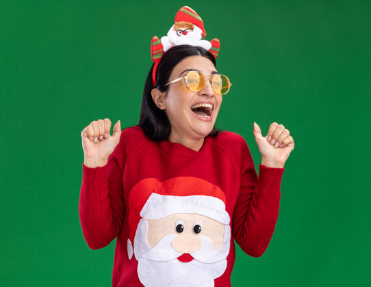 展示兴奋的年轻白人女孩戴着圣诞老人的头带和毛衣 戴着眼镜 竖起大拇指 孤零零地站在绿色的墙上穿着圣诞老人拇指