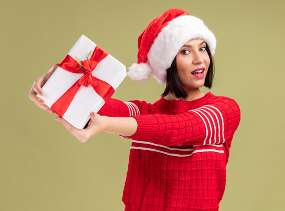 礼物高兴的年轻女孩戴着圣诞帽 向镜头伸出礼包 看着橄榄绿背景上孤立的镜头帽子包装伸展
