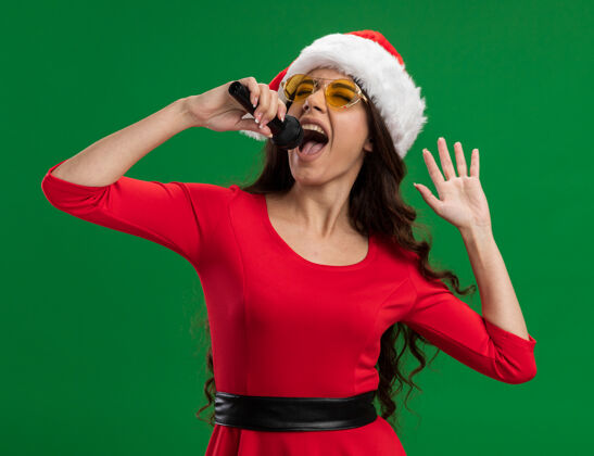 眼睛快乐的年轻漂亮女孩戴着圣诞帽戴着眼镜拿着麦克风手牵手在空中唱歌闭着眼睛隔离在绿色背景上举行快乐关闭