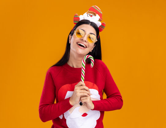 传统快乐的年轻白人女孩戴着圣诞老人的头带和毛衣 戴着眼镜 手持传统的圣诞糖果手杖 垂直地隔离在橙色的墙上 留着复制空间快乐手杖穿着