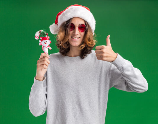 眼镜快乐的年轻人戴着圣诞老人的帽子 戴着红色的眼镜 拿着圣诞糖果手杖 微笑着看着相机 竖起大拇指站在绿色的背景上年轻人糖果抱着