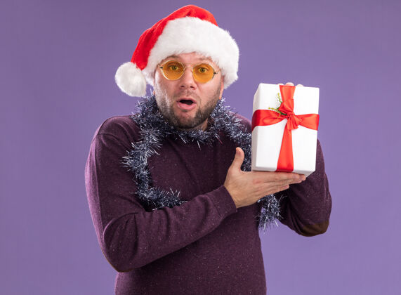 包装印象深刻的中年男子戴着圣诞帽 脖子上戴着金箔花环 戴着眼镜 拿着礼包 看着紫色背景上孤立的相机金属丝帽子眼镜
