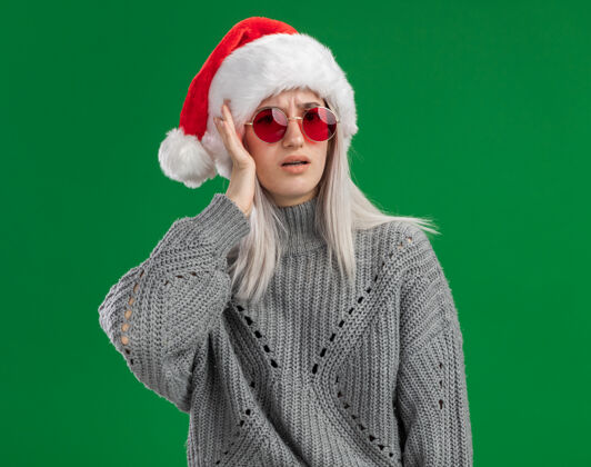 圣诞老人年轻的金发女郎穿着冬季毛衣 戴着圣诞老人帽 戴着红眼镜 一脸困惑 手放在头上 站在绿色的背景上手头帽子