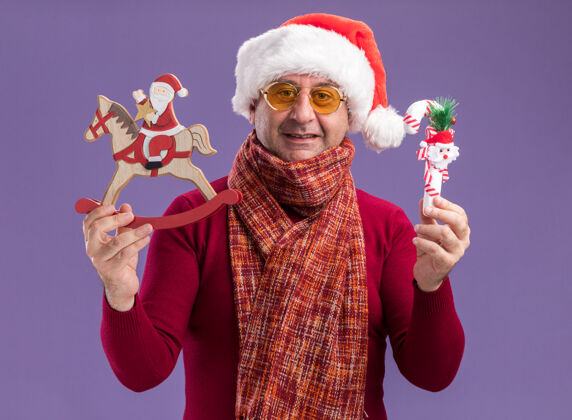 站着中年男子戴着圣诞老人帽 脖子上围着暖和的围巾 戴着黄色眼镜 站在紫色的背景下 展示着圣诞玩具 快乐欢快的微笑年龄帽子周围