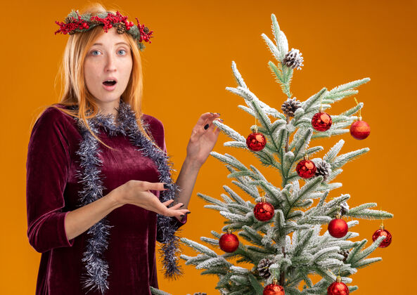 困惑困惑的年轻漂亮女孩站在圣诞树旁边 穿着红色的裙子 脖子上戴着花环 橙色的背景孤立着年轻女孩脖子