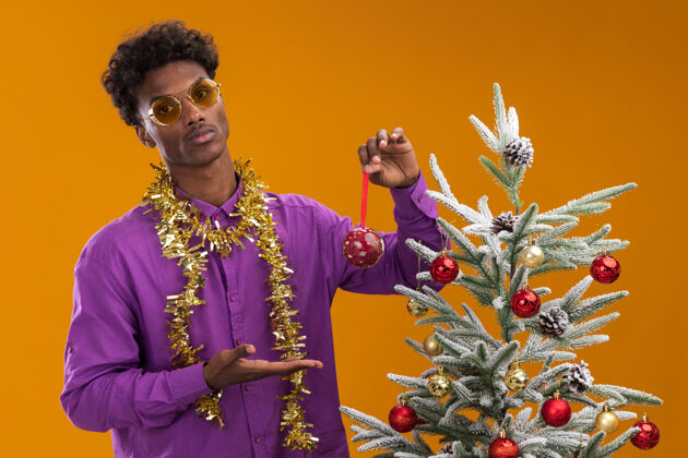 花环自信的美国黑人年轻人戴着眼镜 脖子上戴着金箔花环 站在圣诞树旁 用圣诞饰品装饰圣诞树橙色穿着新