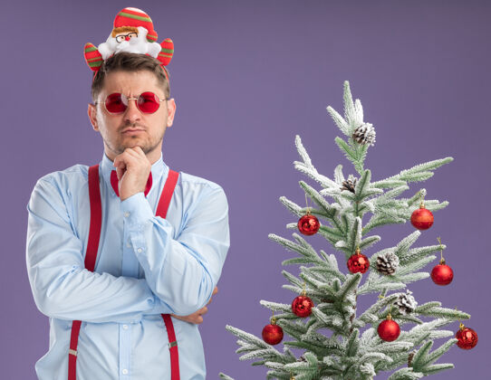 圣诞老人戴着吊带领结的年轻人站在圣诞树旁 戴着圣诞老人和红眼镜 脸上带着沉思的表情看着摄像机 想着紫色的背景脸沉思领带