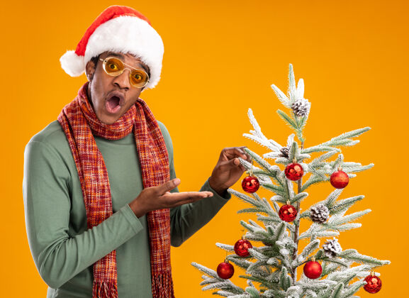 惊讶戴着圣诞帽 脖子上围着围巾的非裔美国人站在圣诞树旁 手臂在橙色背景下显得很惊讶手臂美国围巾