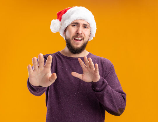 担心穿着紫色毛衣 戴着圣诞老人帽的年轻人看着摄像机焦急地做着防卫的手势 伸出手告诉大家不要靠近站在橙色背景上拿着告诉帽子