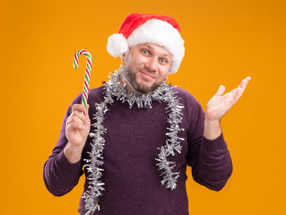 脖子高兴的中年男子戴着圣诞帽 脖子上戴着金箔花环 手里拿着圣诞甜手杖 看着摄像机 镜头里的空手孤立在橙色的背景上花环圣诞周围
