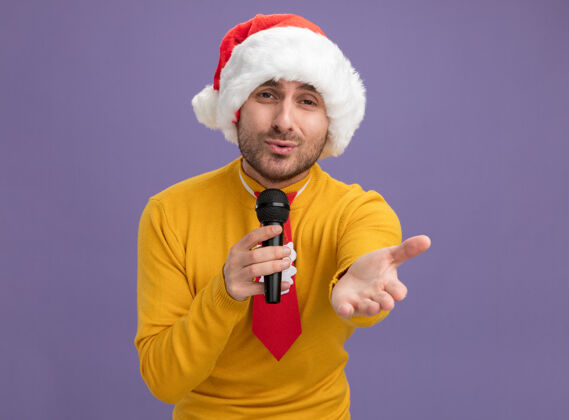 伸展戴着圣诞帽打着领带的白人年轻人拿着麦克风看着摄像机唱歌 伸出手对着隔离在紫色背景上的摄像机圣诞节年轻领带