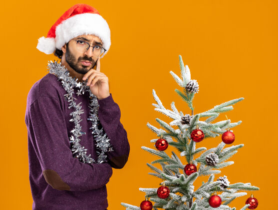 脸颊想着站在圣诞树旁的年轻帅哥 戴着圣诞帽 脖子上戴着花环 手指放在脸颊上 孤立在橙色的背景上帽子树脖子