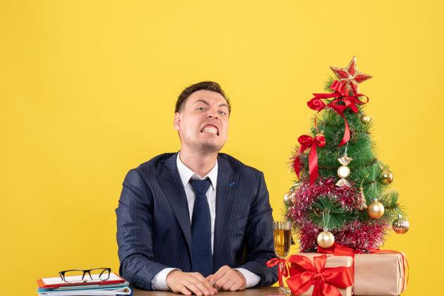 办公室愤怒的男人坐在圣诞树旁的桌子前 在黄色的墙上复制礼物的地方礼物坐着会议
