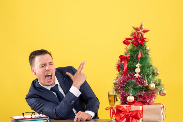 坐前视图愤怒的男子 使手指枪的标志坐在圣诞树和礼物附近的黄色墙上的桌子上手指商人制造