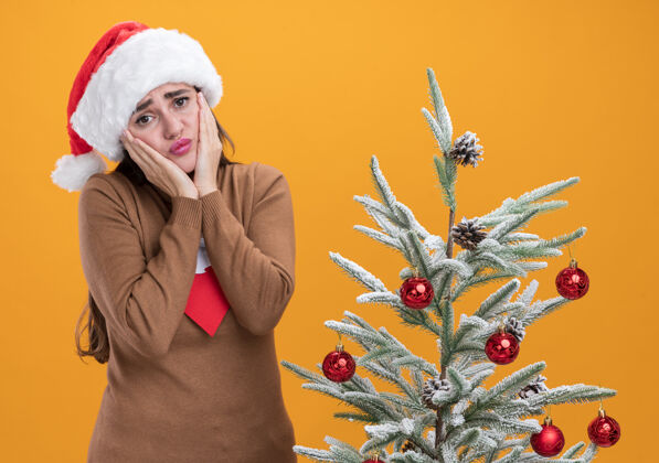 身边戴着圣诞帽打着领带的悲伤的年轻漂亮女孩站在圣诞树旁 手放在橘色背景上孤立的脸颊上悲伤放着帽子
