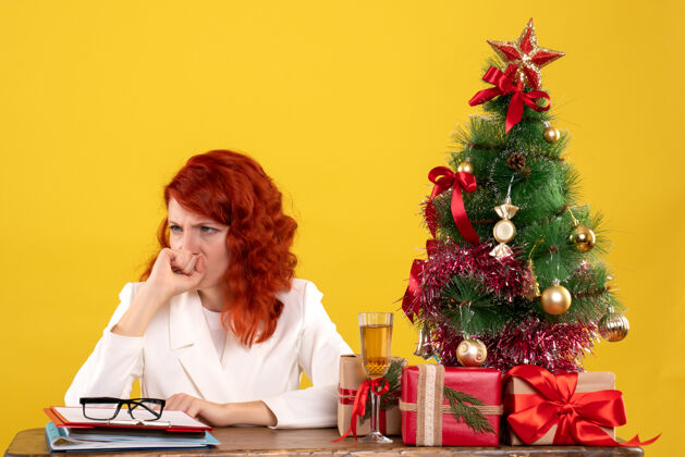圣诞节女工人坐在桌子后面 手里拿着圣诞礼物和黄色的圣诞树办公室树人