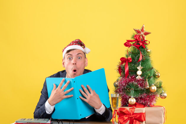 圣诞节令人惊叹的商人坐在圣诞树旁的桌子前 黄色的墙上摆着礼物人微笑礼物