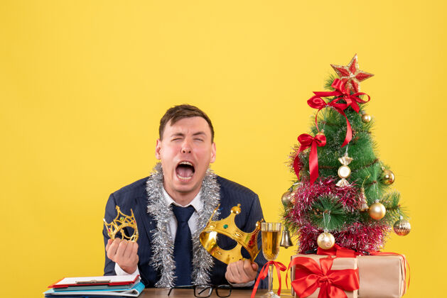 哭着商务人士坐在圣诞树旁的桌子前哭泣 黄色墙上摆着礼物圣诞节生意人生意