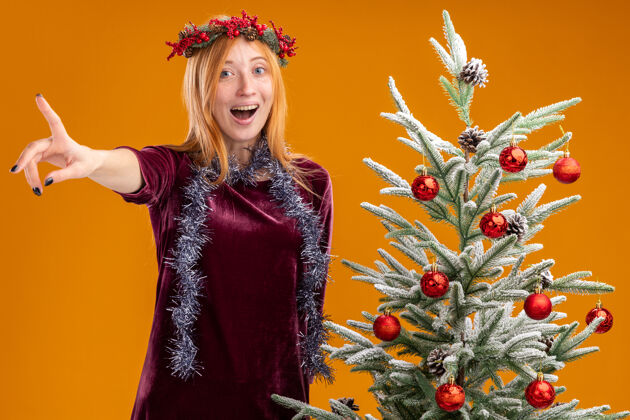 花环站在圣诞树旁微笑的年轻漂亮女孩 身穿红色连衣裙 脖子上戴着花环 镜头被隔离在橙色背景下点衣服树