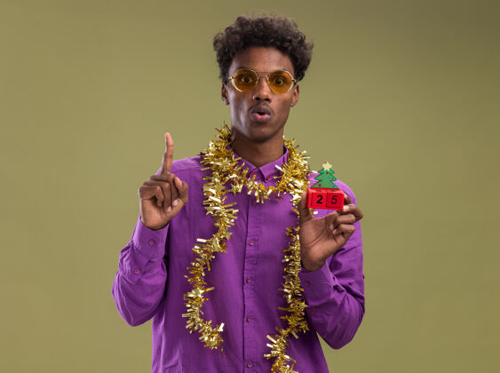 树令人印象深刻的年轻非裔美国男子戴着眼镜 脖子上戴着金属丝花环 手里拿着圣诞树玩具和日期 看着相机指着橄榄绿背景上孤立的复制空间圣诞节日期年轻