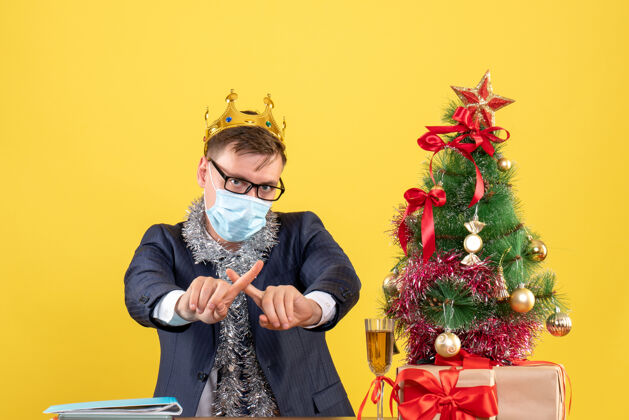 正常商务人士交叉手指坐在圣诞树旁的桌子前 黄色墙壁上的礼物领结领带树