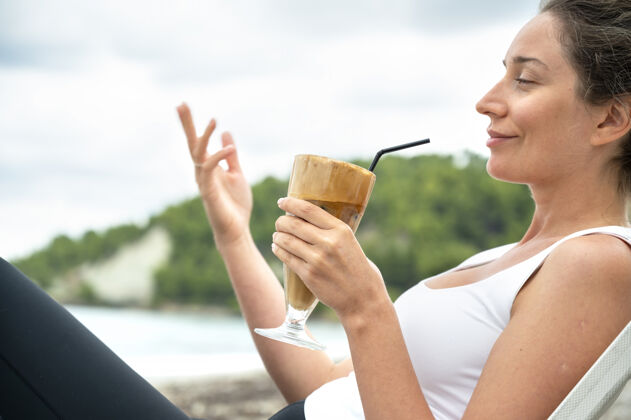 周到微笑的高加索妇女拿着咖啡饮料在沙滩上与泡沫和喝稻草与山侧举行蓝色