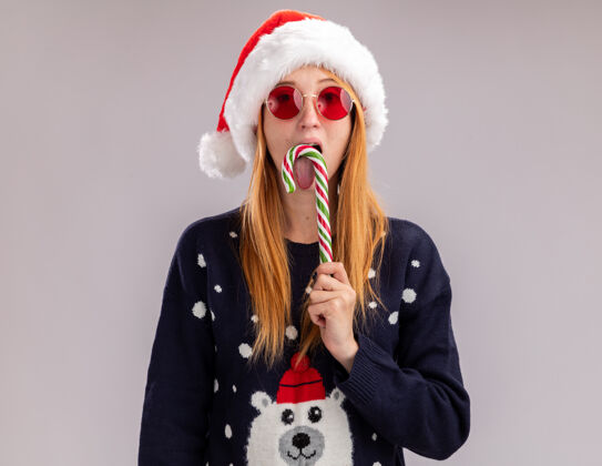 穿年轻漂亮的女孩戴着圣诞帽 戴着眼镜 抱着和舔着隔离在白墙上的圣诞糖果糖果舔圣诞节
