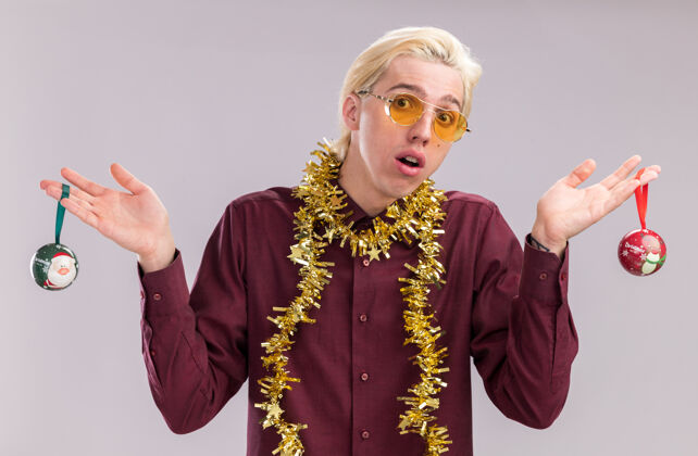 圣诞节笨手笨脚的年轻金发男子戴着眼镜 脖子上戴着金属丝花环 手里拿着圣诞饰品 看着隔离在白色背景上的相机眼镜脖子金属丝