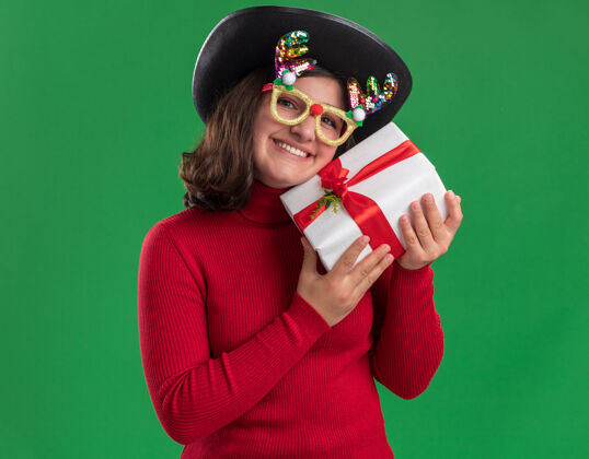 开心穿着红毛衣 戴着滑稽眼镜 戴着黑帽子 拿着礼物的快乐小女孩站在绿色的背景下看着镜头 开心地笑着礼物抱着微笑