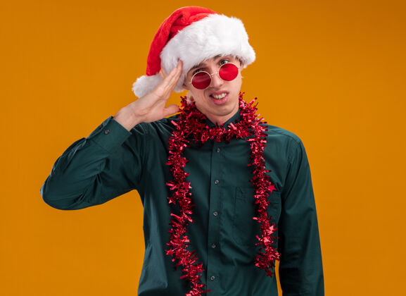 触摸困惑的年轻金发男子戴着圣诞帽 戴着眼镜 脖子上戴着金属丝花环 看着相机触摸着孤立在橙色背景下的头头脖子困惑