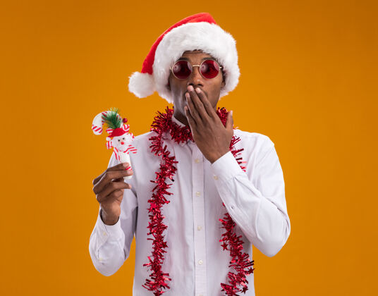 圣诞老人关心的美国黑人年轻人戴着圣诞帽 戴着眼镜 脖子上戴着金箔花环 手里拿着糖果手杖 看着相机 手放在嘴巴上 在橙色背景下与世隔绝装饰品非裔美国人嘴